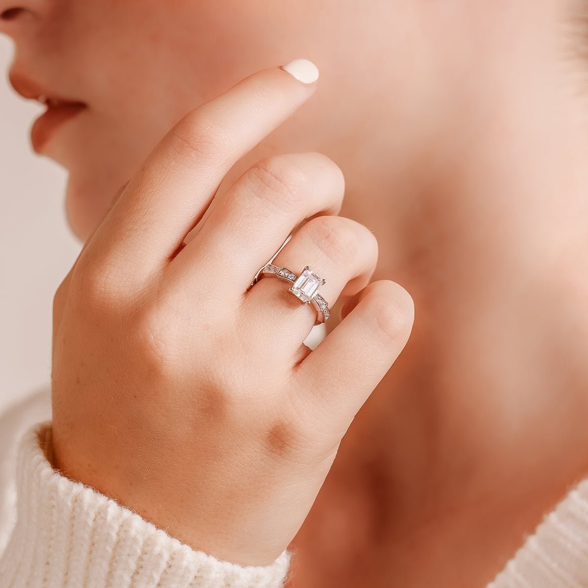 Close-up of a hand near a face, fingertip with cream and a silver-toned ring with emerald-cut center stone.