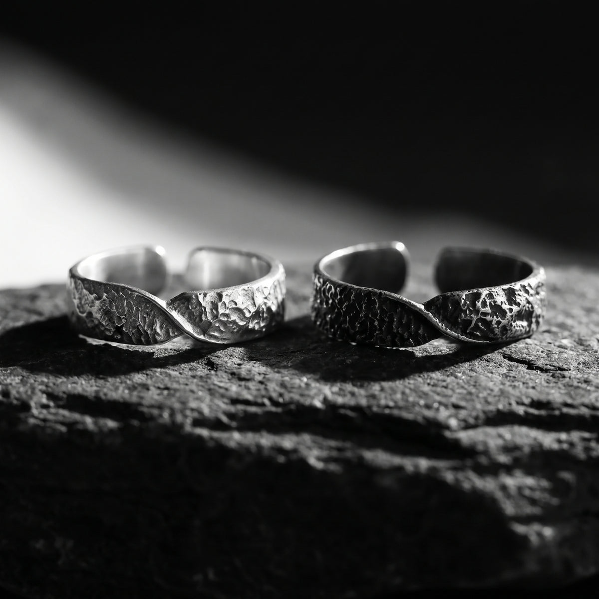 Two textured silver twist rings in natural and dark oxidized finishes resting side by side on dark stone.