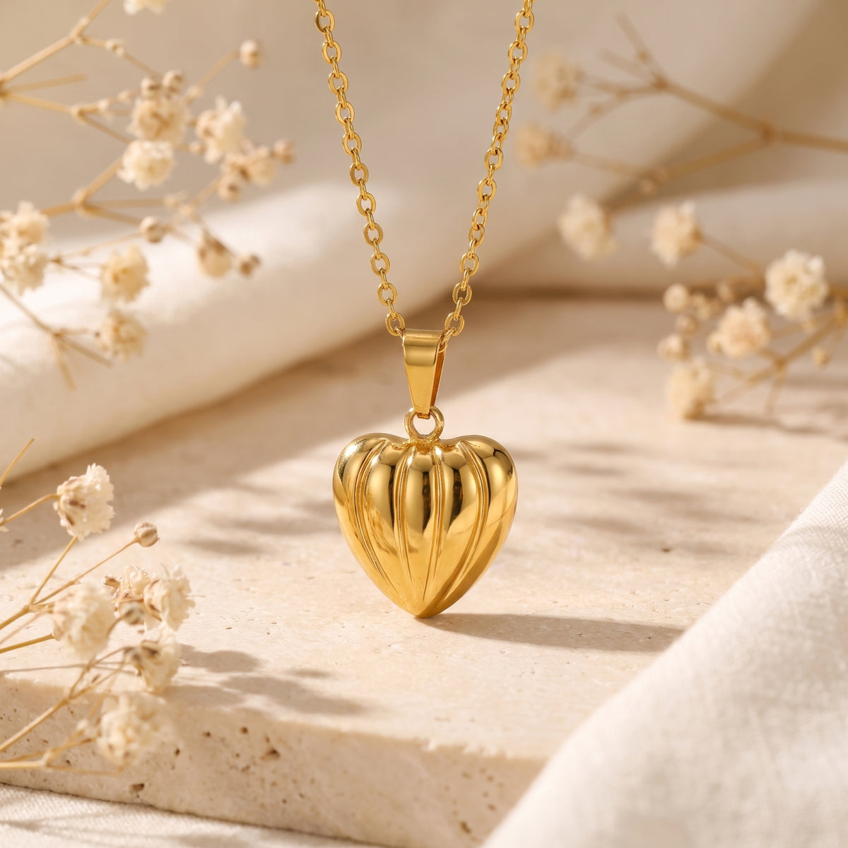 Front closeup of a gold-tone ribbed heart pendant on a fine cable chain, set on stone among dried white flowers.
