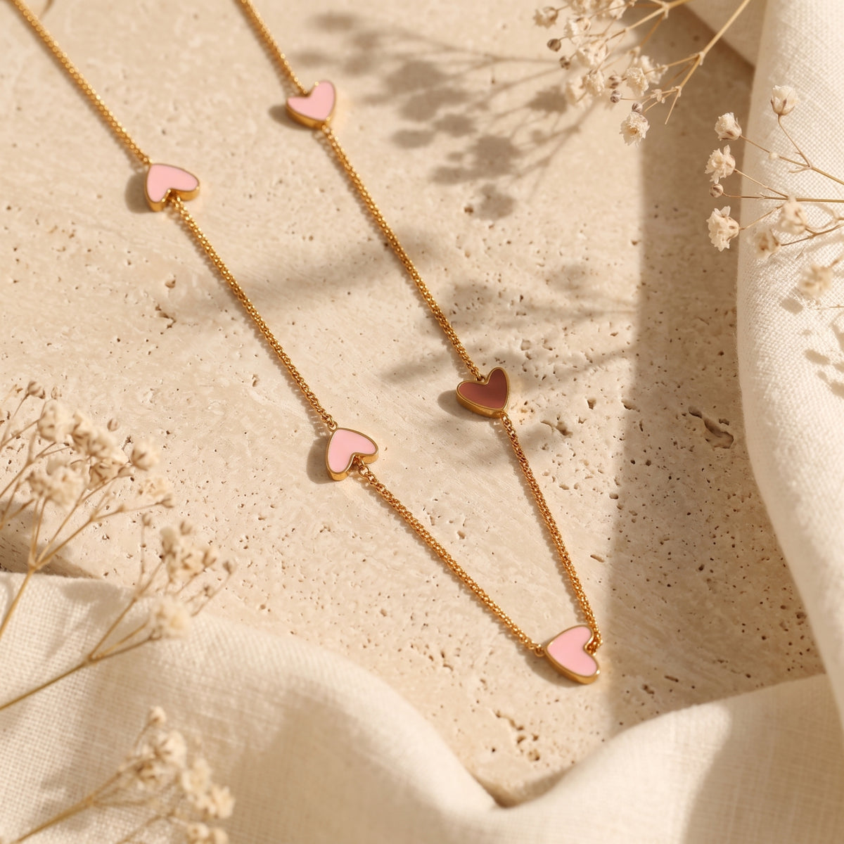 Gold-tone station necklace with pink and rose enamel heart charms on a textured stone surface with dried florals.