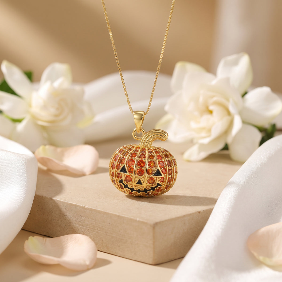Gold-tone pumpkin pendant necklace displayed on a beige platform among white flowers and draped white fabric.