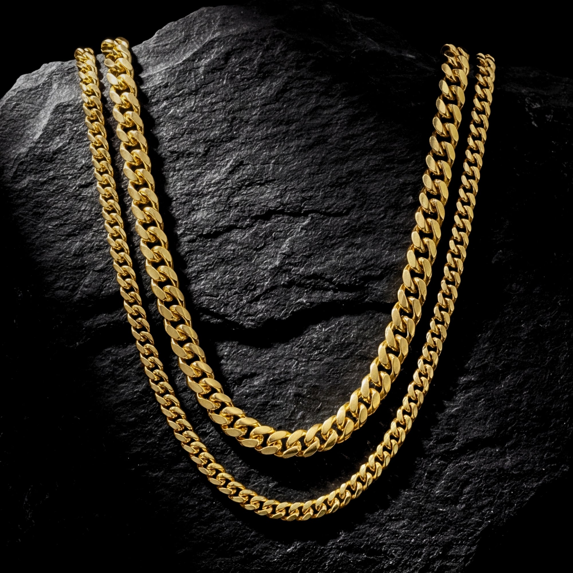 Two gold Cuban link stainless steel chains displayed on a dark textured rock.