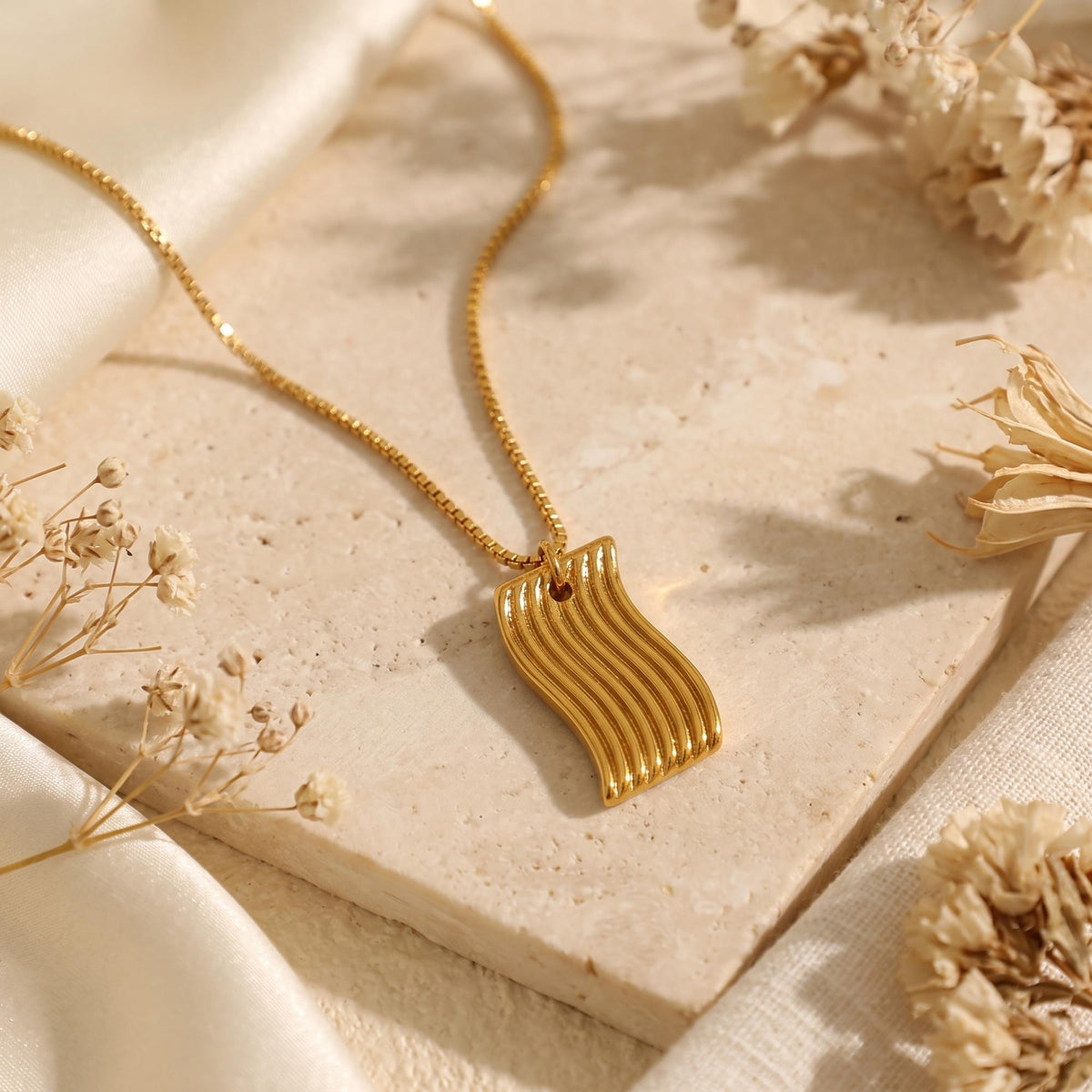Gold-tone wavy ridged pendant necklace displayed flat on a beige stone tile alongside white silk and dried flowers.