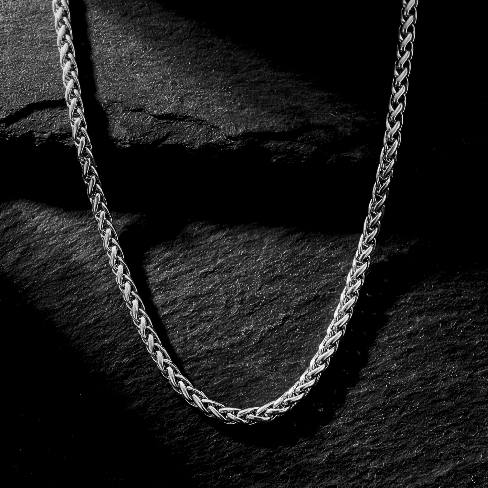 Silver stainless steel wheat chain necklace draped over a dark, textured stone background.