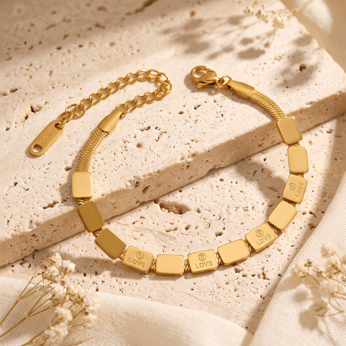 Gold-tone link bracelet with engraved tiles and snake chain section on a textured stone block.