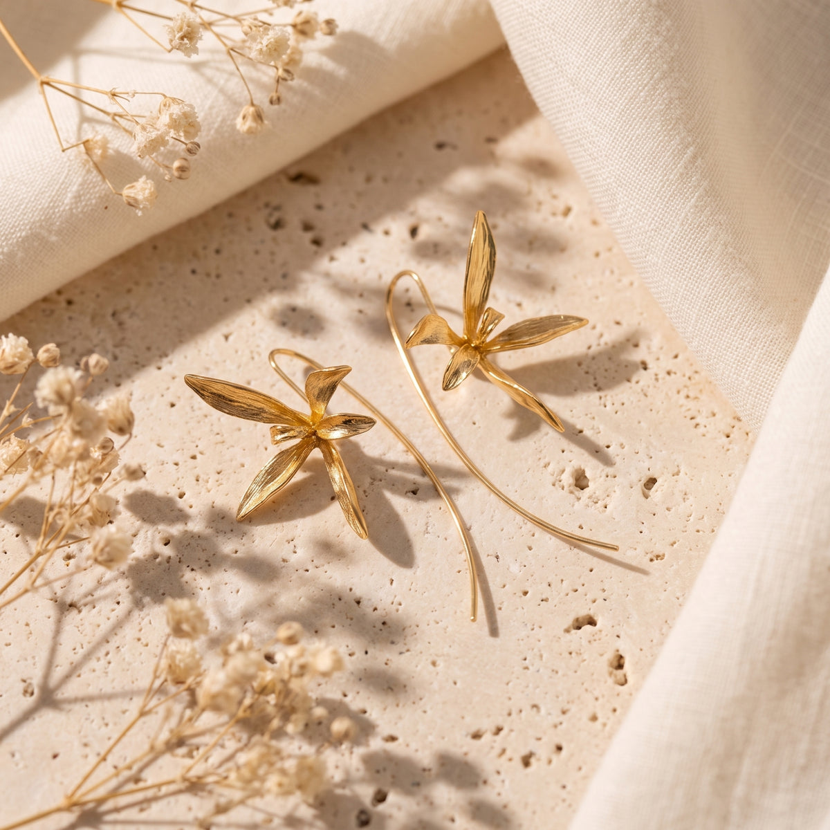 Pair of gold-finish orchid drop earrings displayed on a textured beige stone slab beside linen fabric.