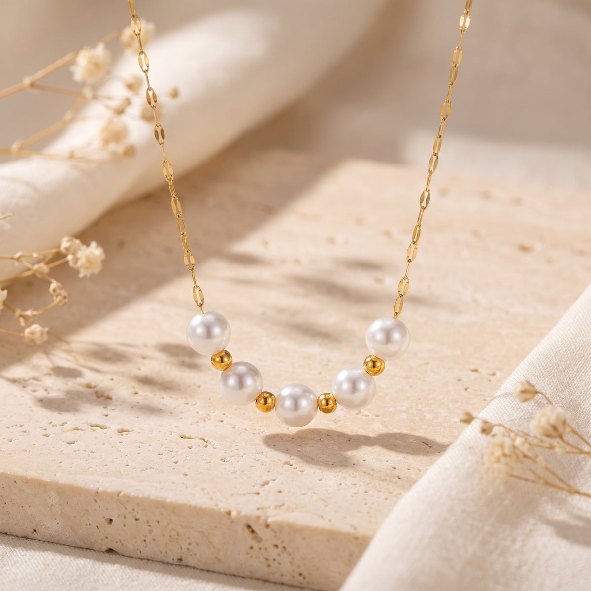 Gold-tone chain necklace with white simulated pearls and polished gold beads, suspended over a beige travertine slab.