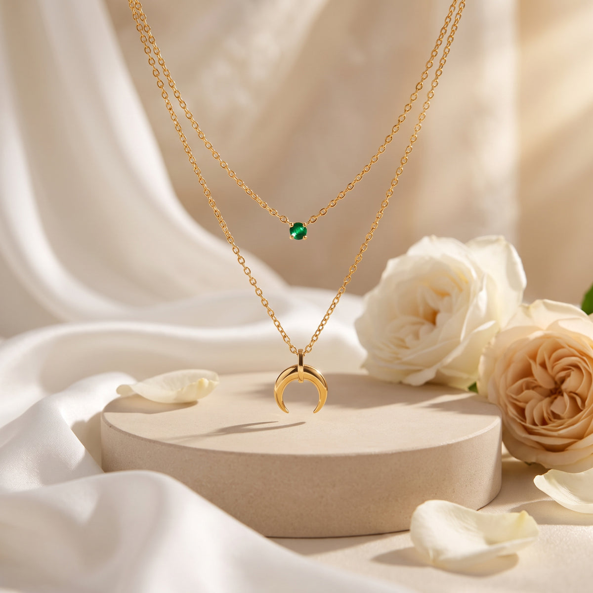 Gold-tone layered necklace with green cubic zirconia and crescent pendant displayed on a beige pedestal among white roses.