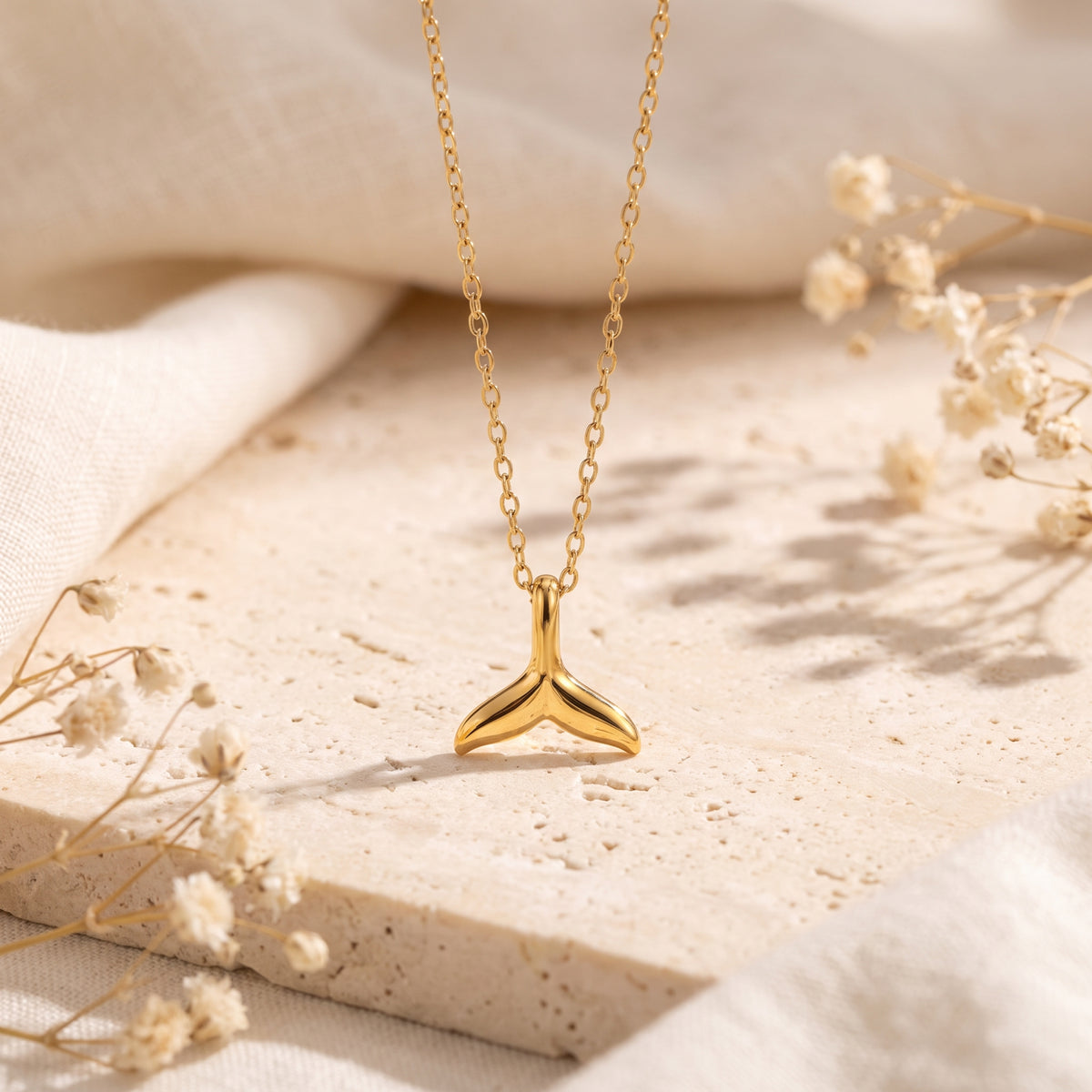Gold-tone whale-tail pendant necklace hanging with its pendant resting on a pale stone slab among dried flowers.
