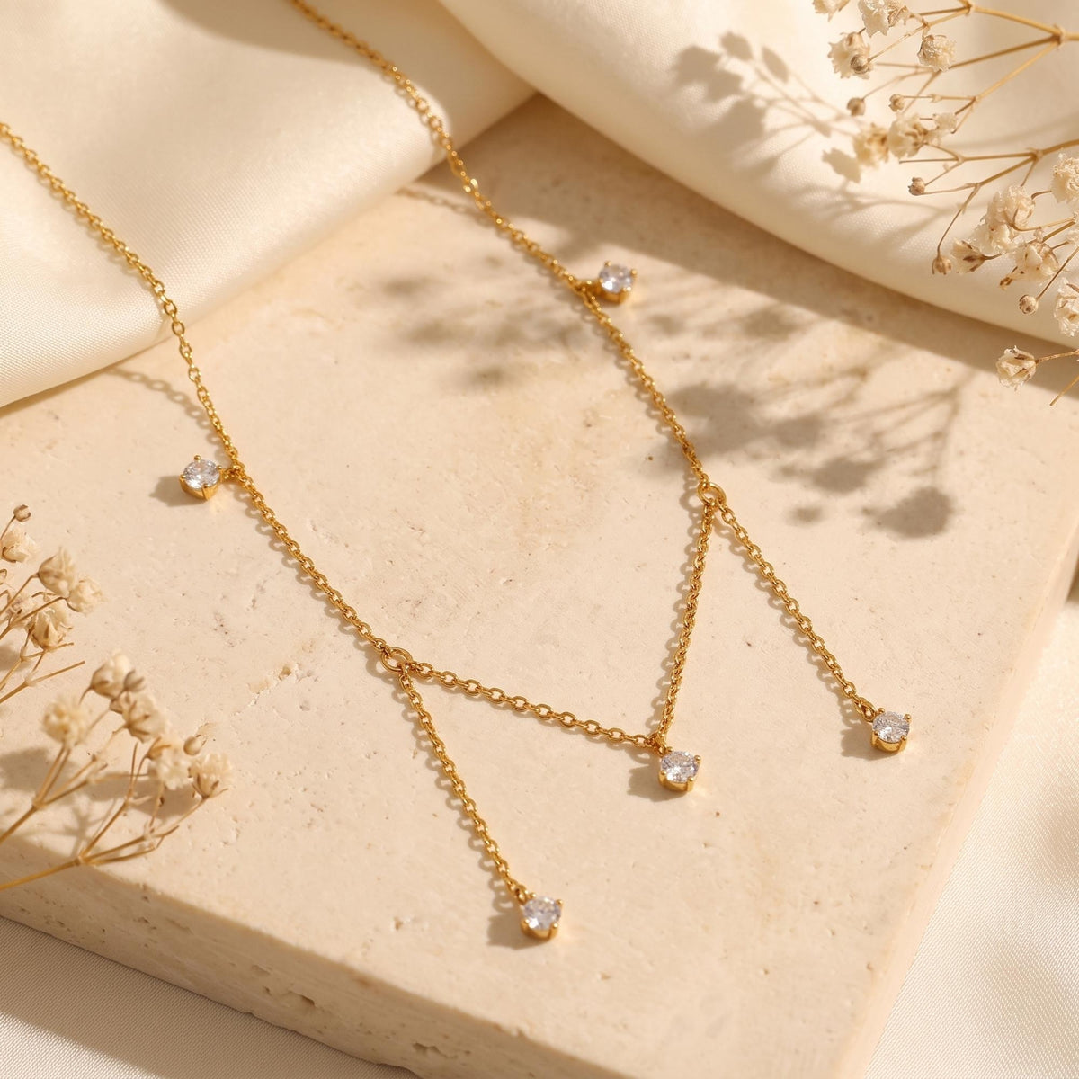 Gold-tone drop necklace with five clear stones displayed on a beige stone slab with dried flowers.