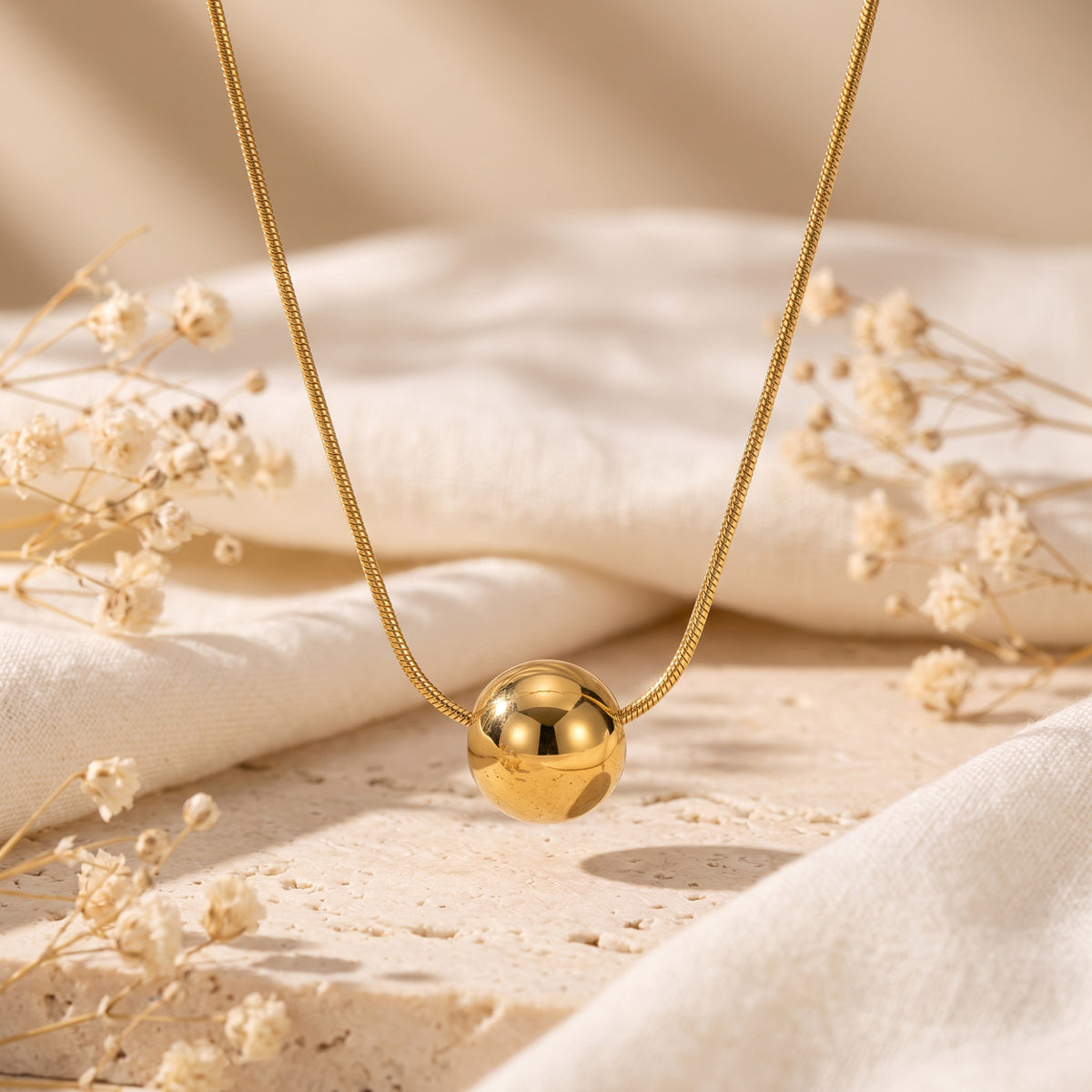 Gold-tone sphere necklace on linen and textured stone, surrounded by dried white flowers.