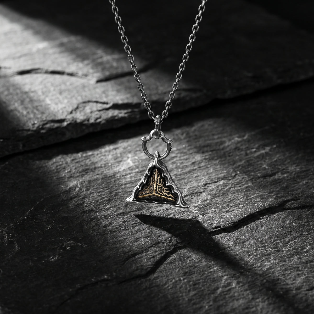 Silver triangular pendant necklace with a gold-toned eye motif hanging over a dark slate surface.