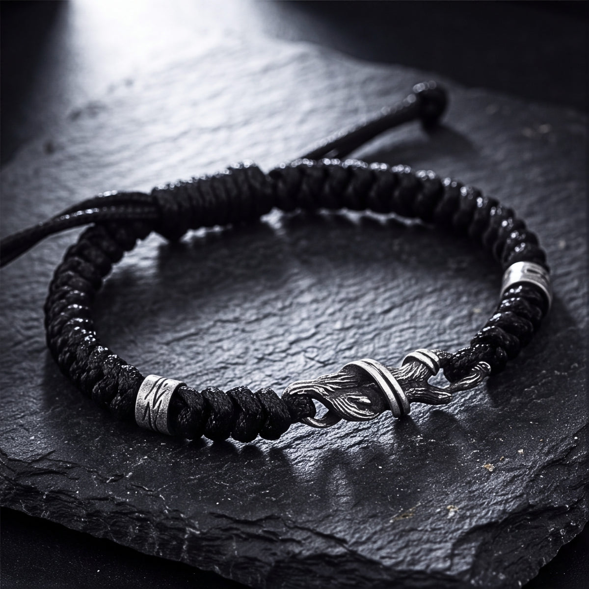 Black woven cord bracelet with a branch-textured silver hook clasp and two silver bands, arranged on a slate surface.