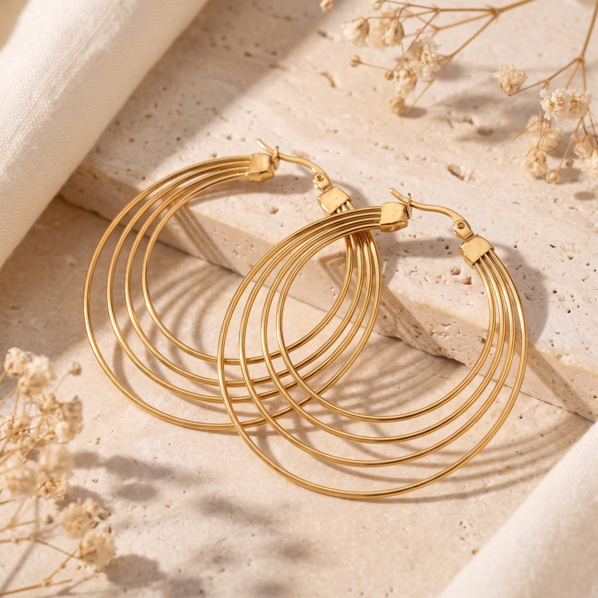 Pair of gold-tone four-ring hoop earrings resting on textured stone with dried flowers and fabric.