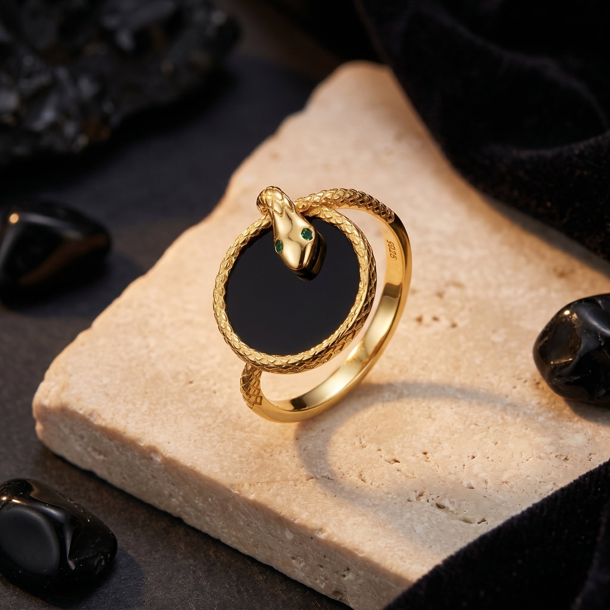 The Regalia gold plated snake statement ring with round black onyx and green eyes on a stone slab.