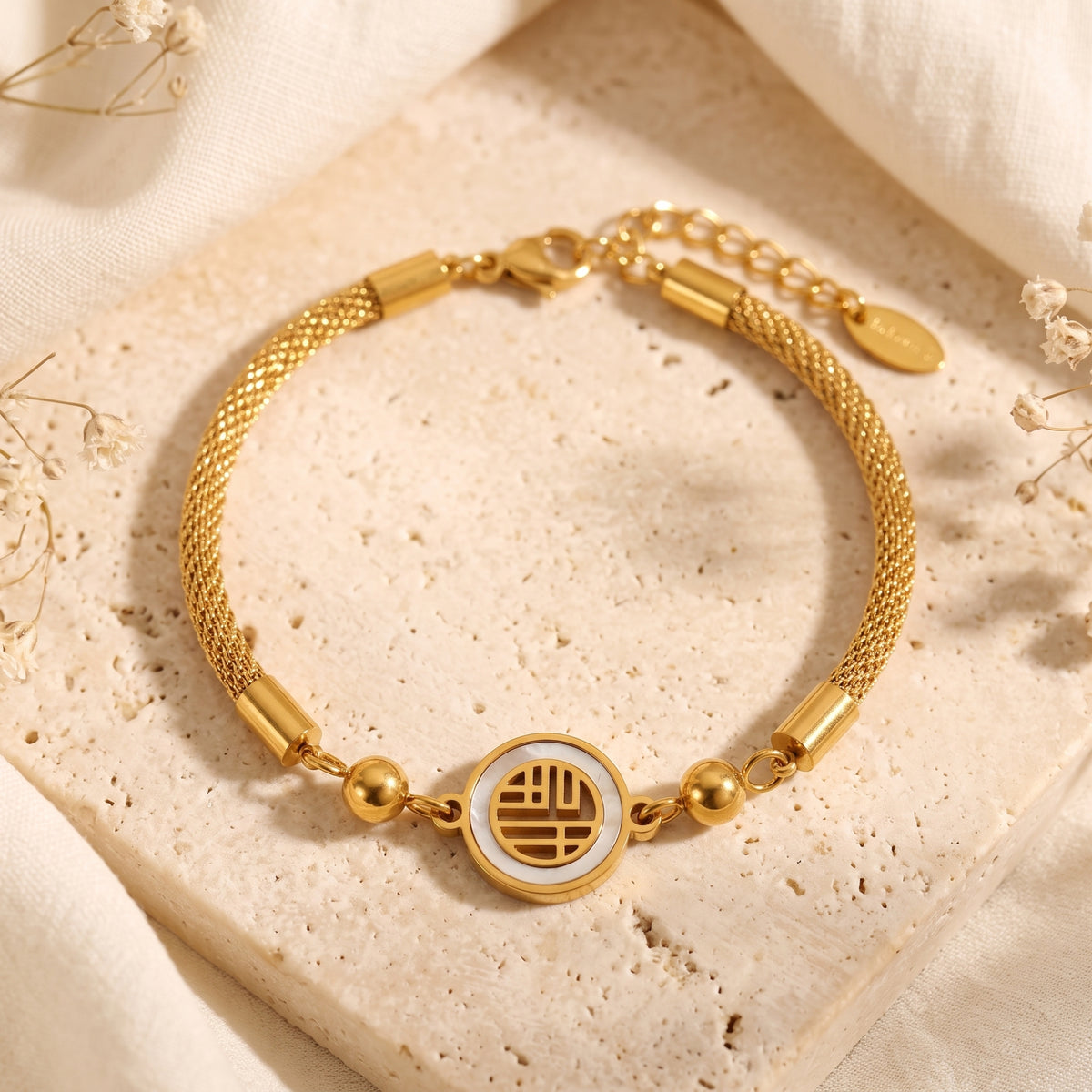 Gold-tone mesh bracelet with a central mother-of-pearl symbol medallion, laid flat on a stone display.