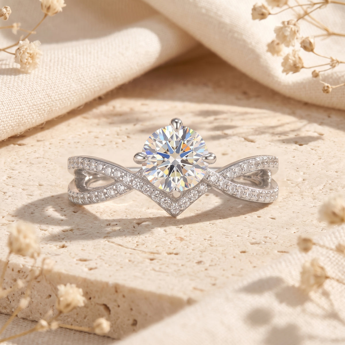Front view of a silver ring with a round center stone, pave V-cradle, and crisscross infinity band on a stone surface.
