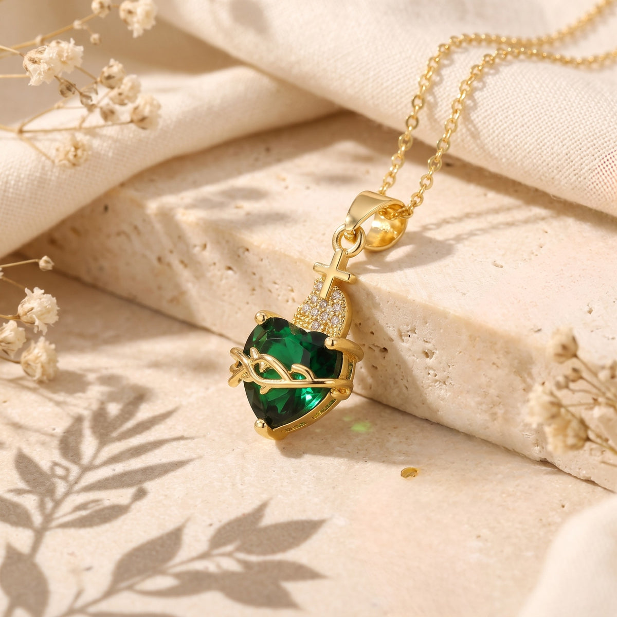 Gold-tone sacred heart pendant with a green crystal, thorn vine, and cross on a beige stone block.
