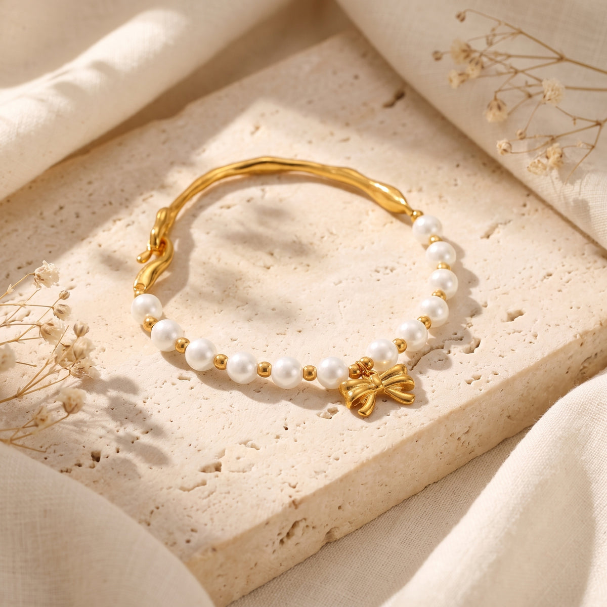 Overhead view of a pearl bracelet with a gold-tone bow charm and hook clasp on a beige stone slab.