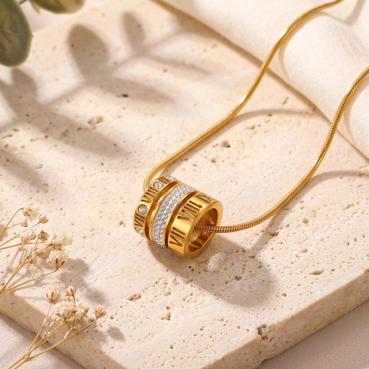 Gold-toned Roman numeral barrel pendant necklace on a textured stone surface with dried flowers.