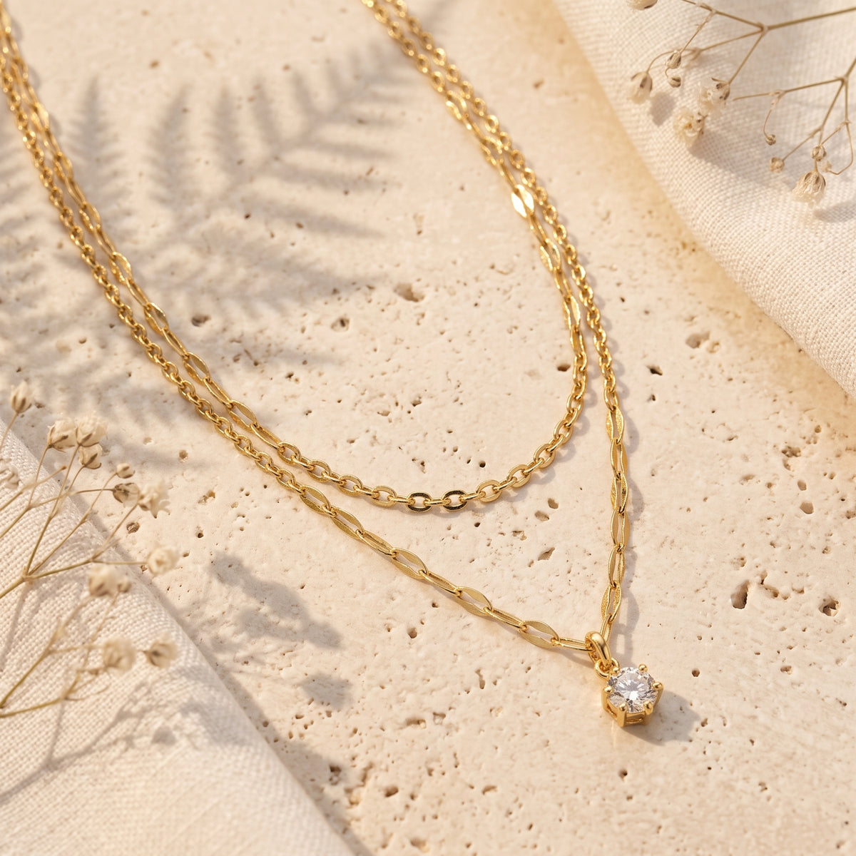Gold-tone double-strand necklace with a round gem pendant laid flat on a beige stone surface.