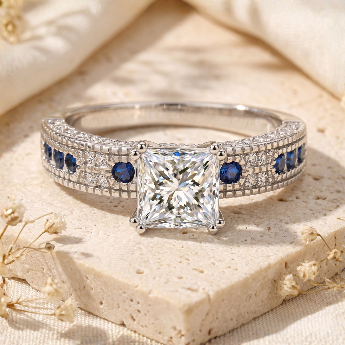 Princess-cut CZ silver ring with blue sapphire accents on a stone surface.