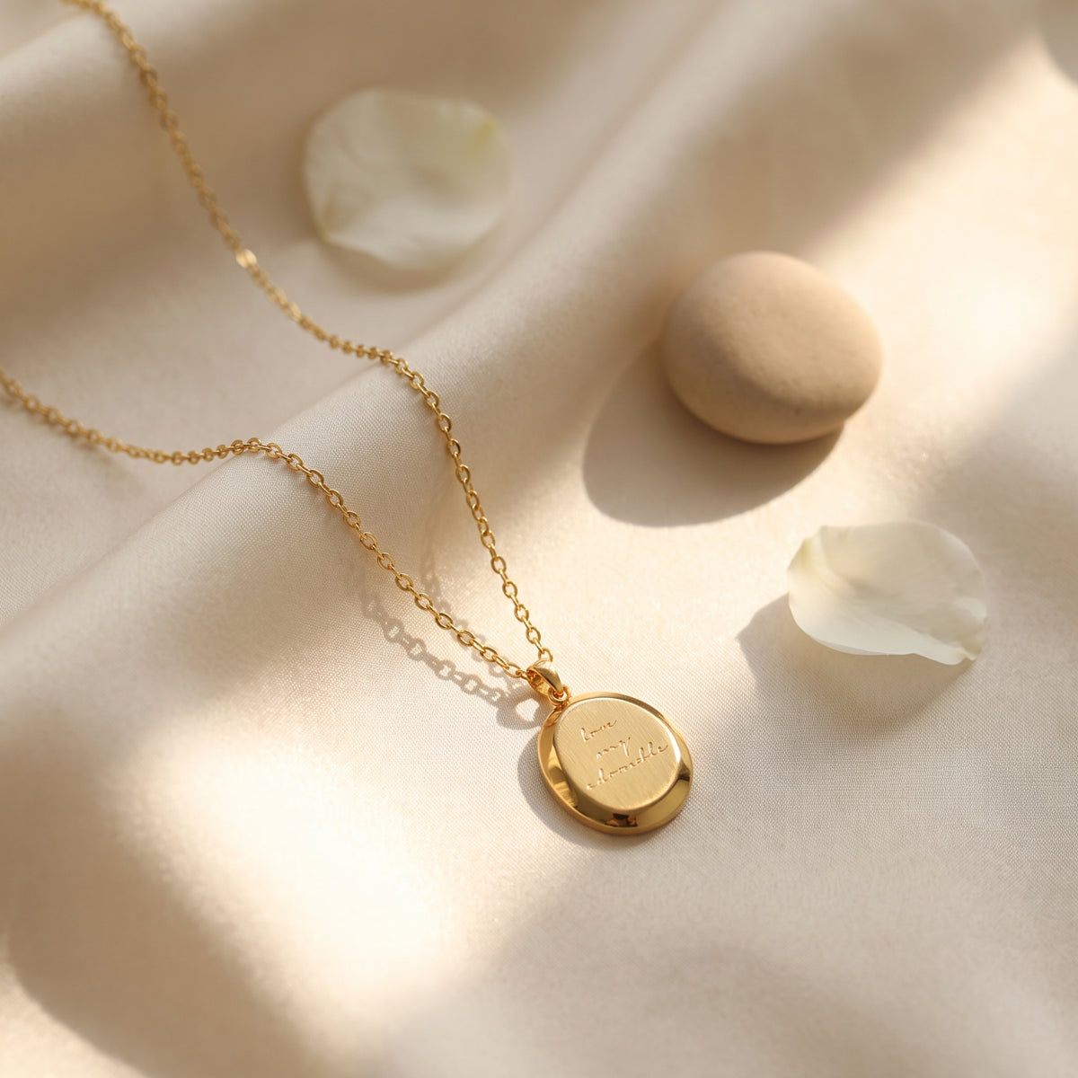 Gold-tone round medallion necklace with cursive engraving, laid on draped cream fabric with stones and a white petal.