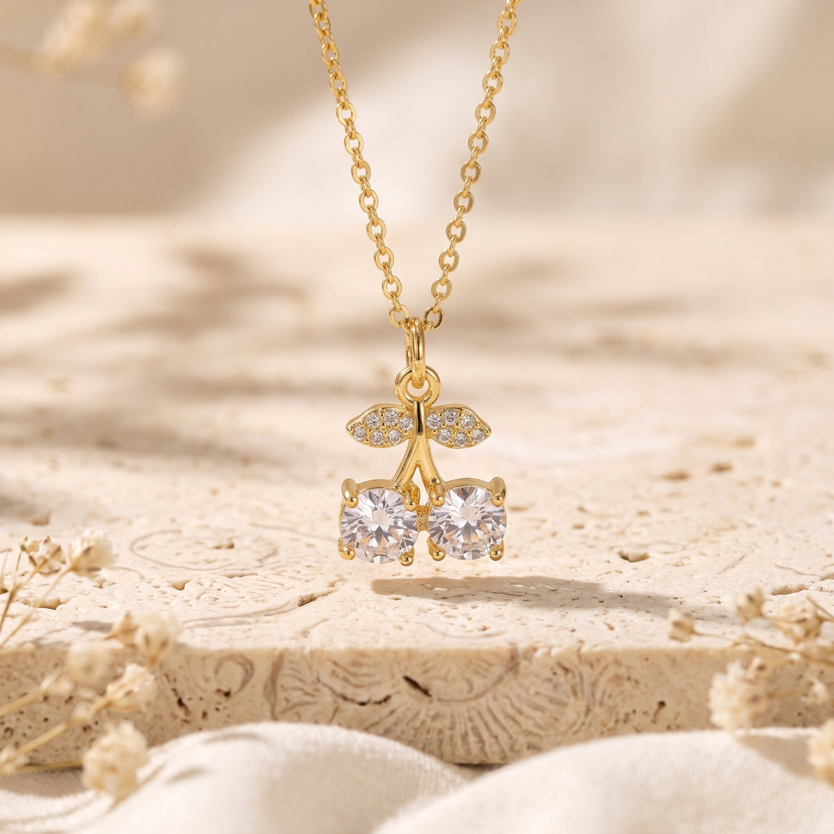Gold-tone cherry pendant necklace with twin round clear stones and pavé leaves suspended over a stone base.