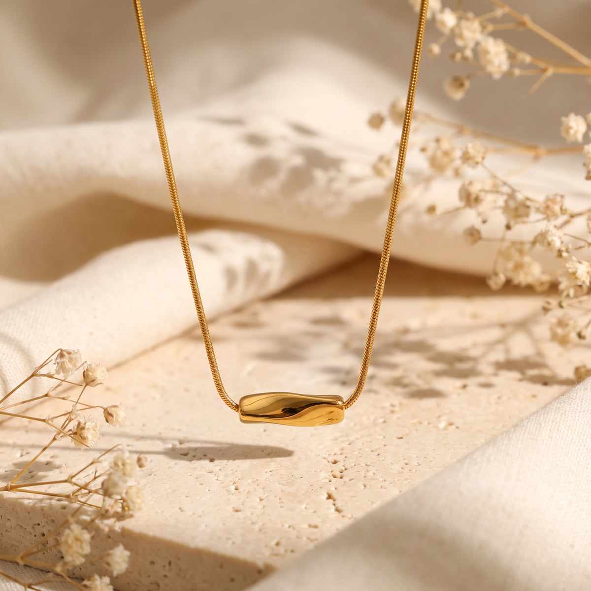 Close-up of a gold-tone wave slide pendant on a snake chain over beige stone.
