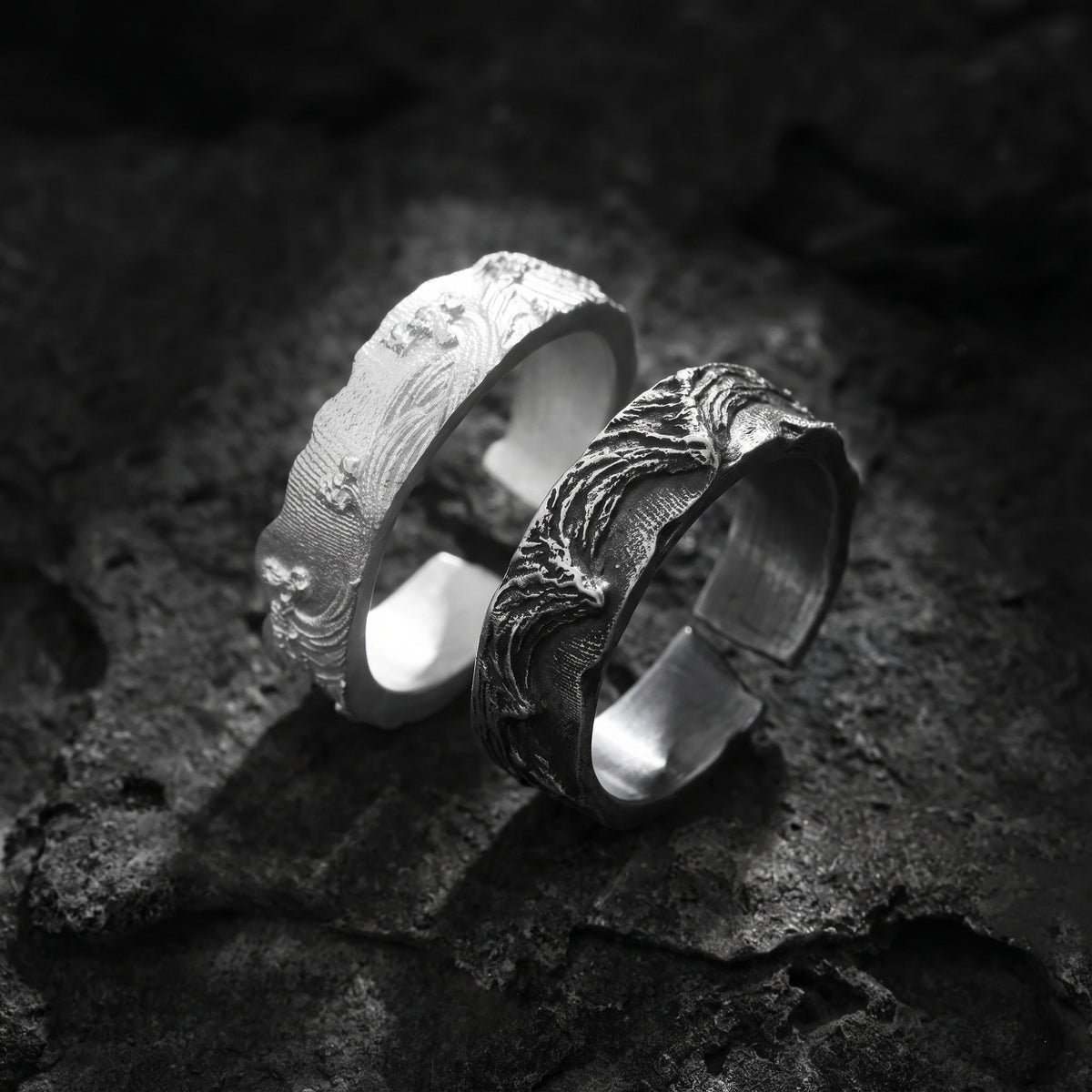 Close-up of dark-toned and bright silver wave open-band rings arranged on rough rock.