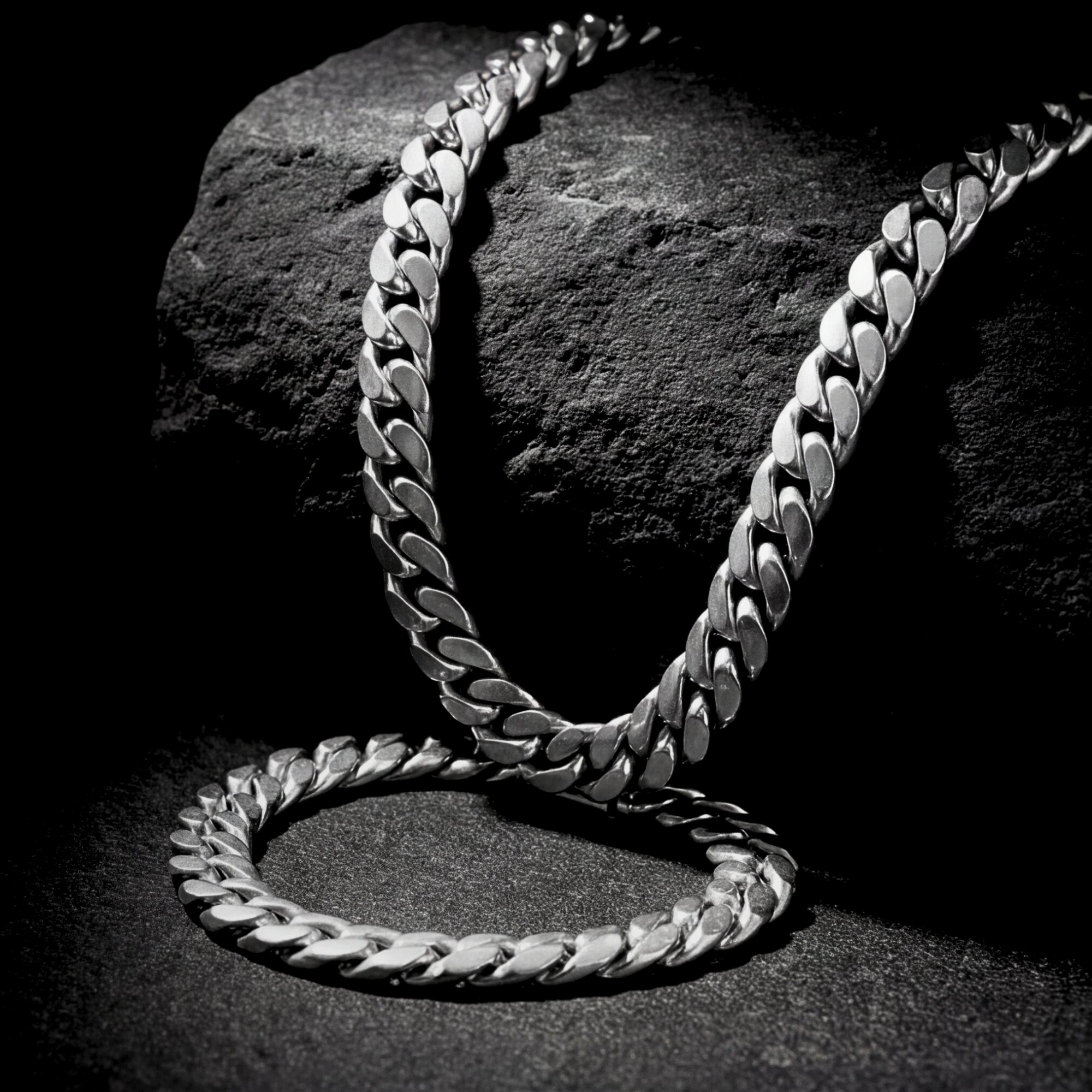 Silver stainless steel Cuban link chain and bracelet set draped over a dark textured rock.