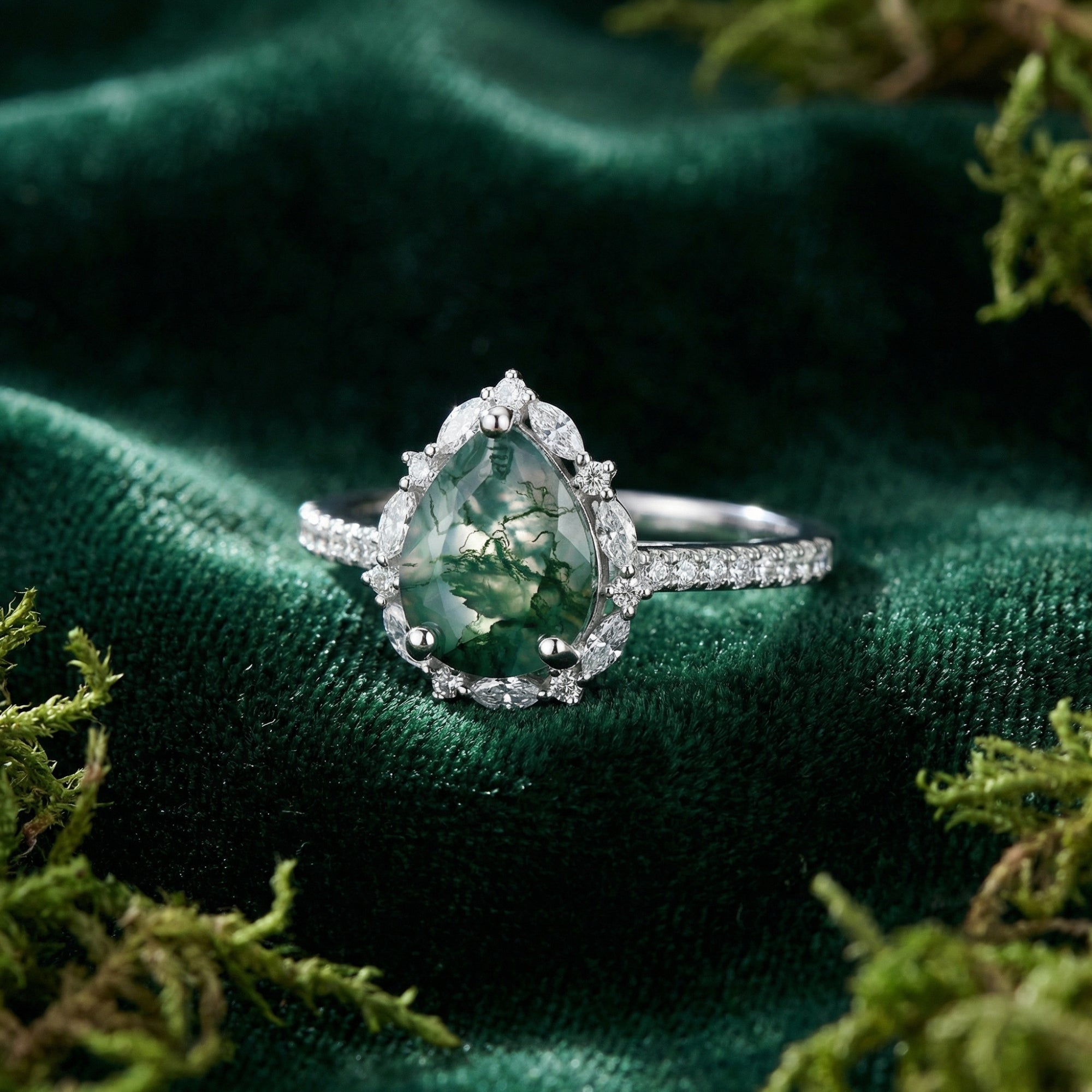 Pear cut moss agate ring in S925 sterling silver with a marquise diamond halo, displayed on green velvet with moss.