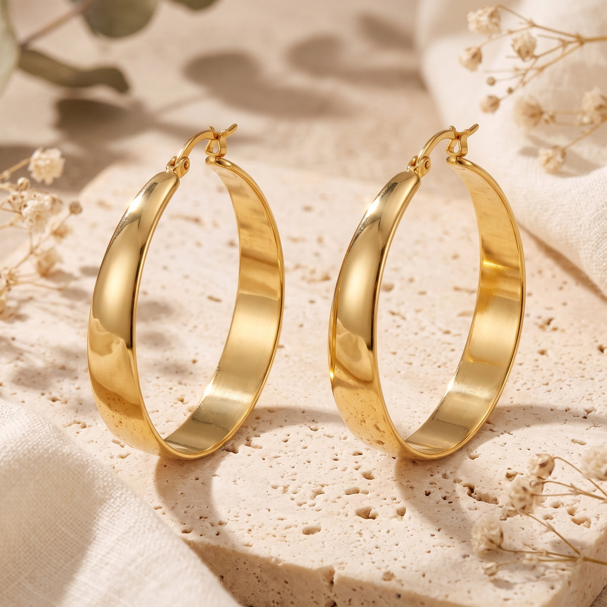 Pair of gold-tone tapered hoop earrings standing upright on a textured stone surface with dried flowers.