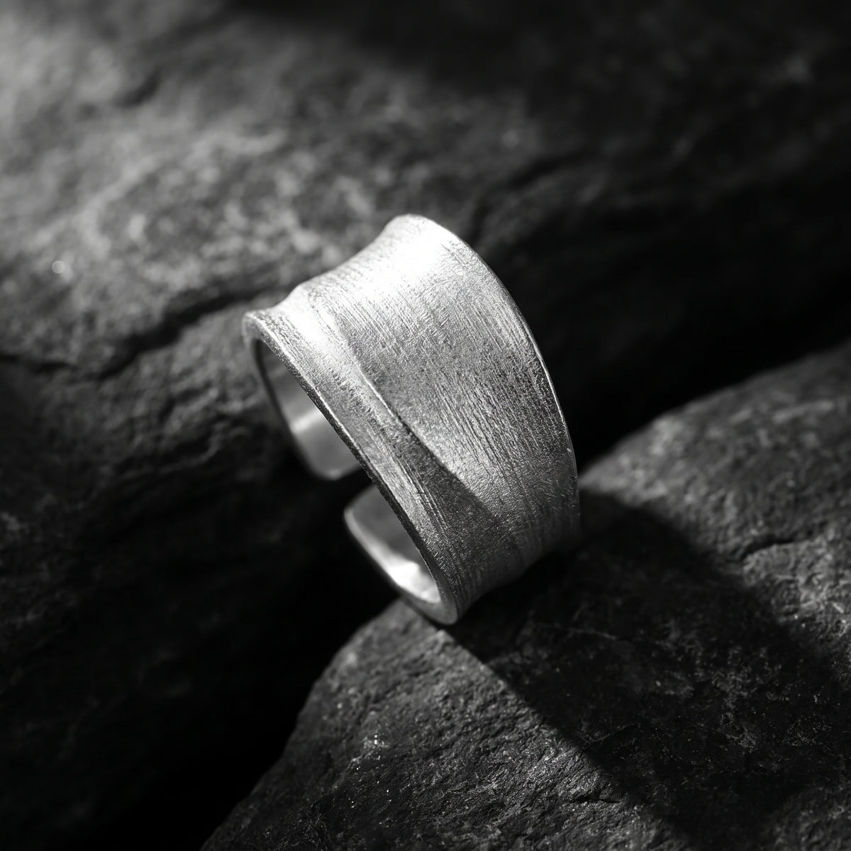 Angled close-up of a wide brushed silver open ring resting on dark rock.