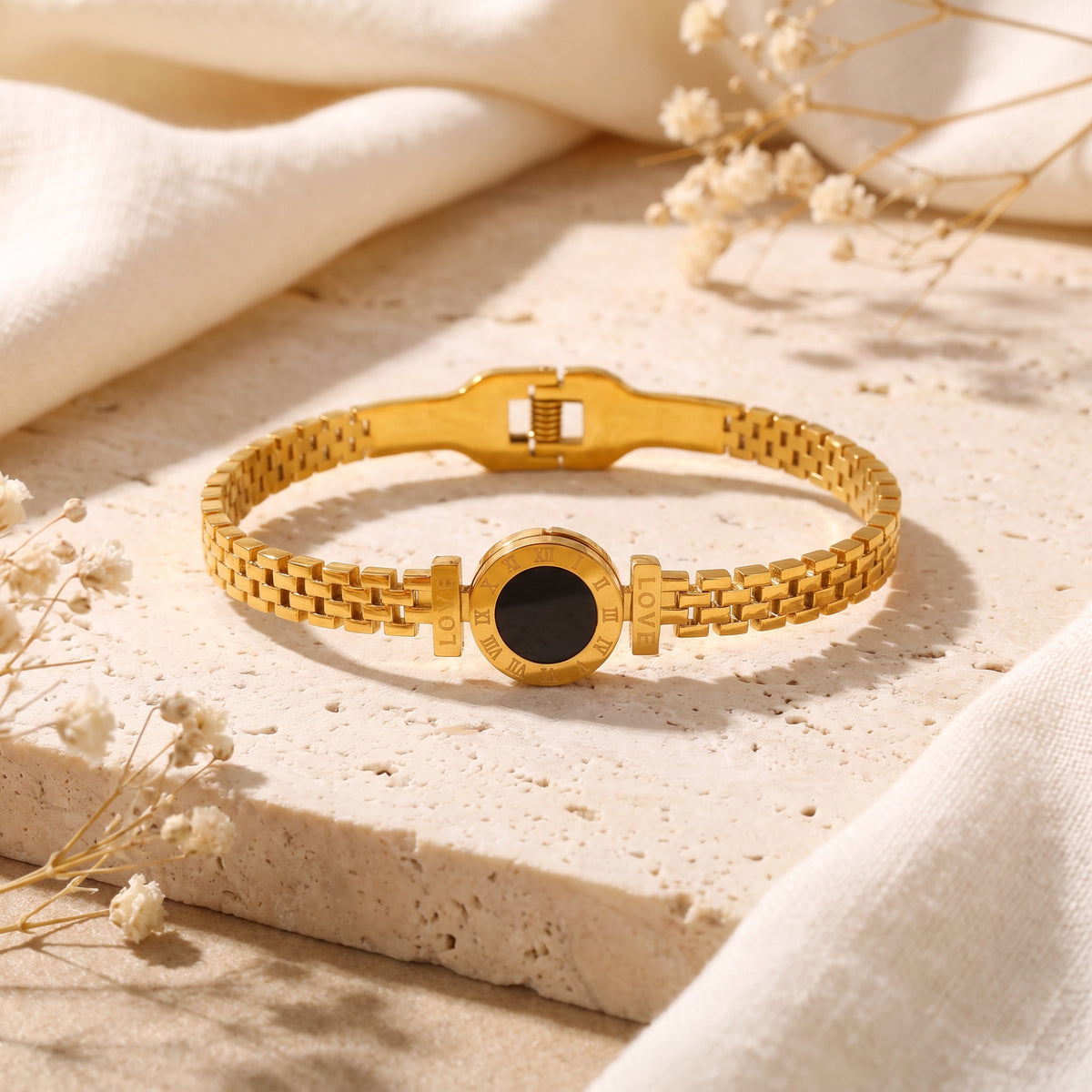 Gold-tone link bracelet with a black circular Roman numeral medallion, resting on a stone surface.