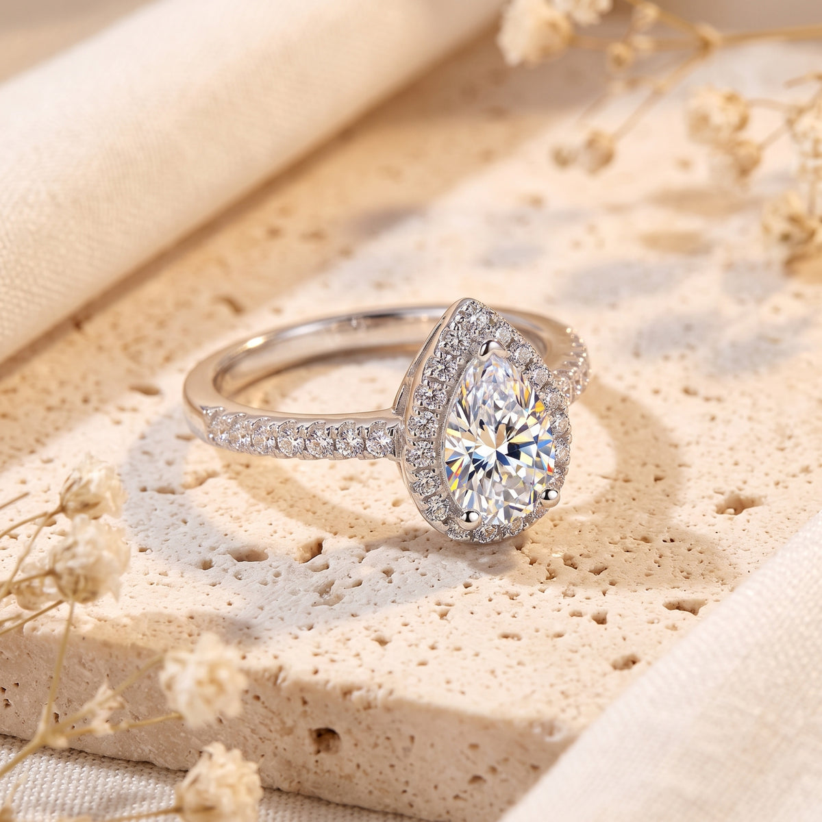 Silver pear-cut halo ring with a pave band displayed on a beige stone surface with dried flowers.