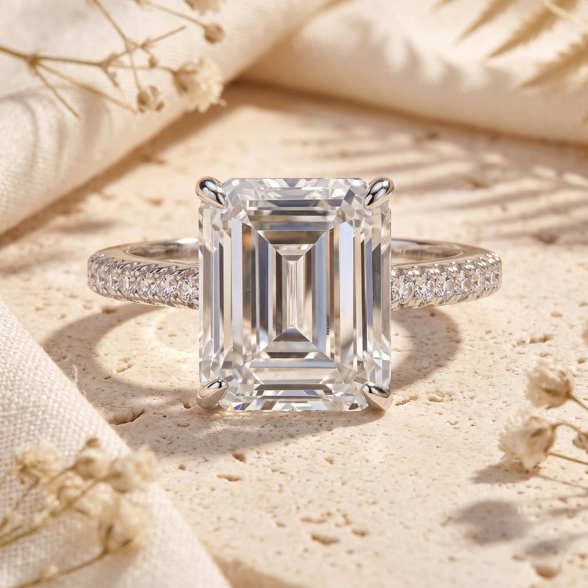 Front view of an emerald-cut solitaire ring with pave accent stones on a silver band, set on textured stone.