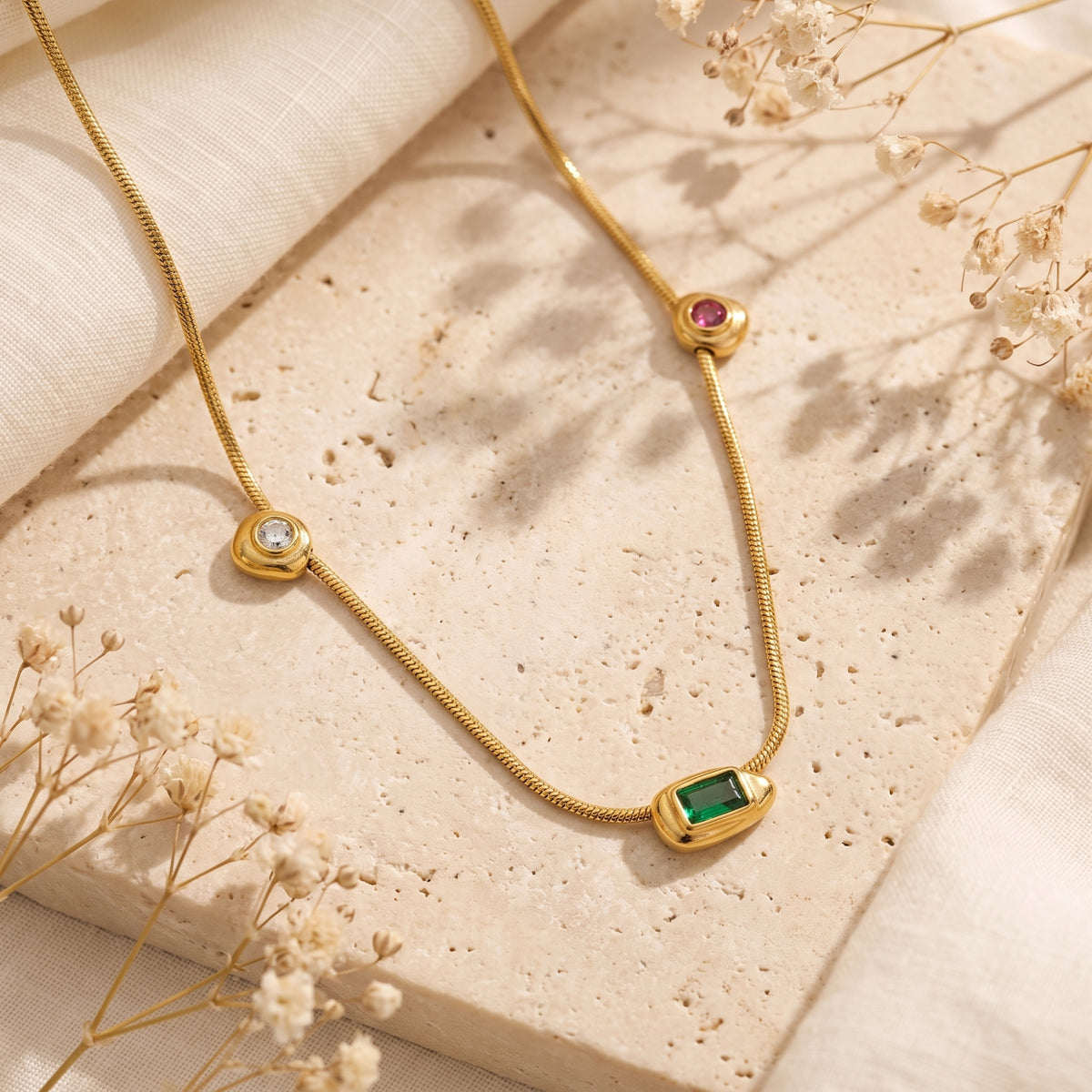 Gold station necklace with green, clear, and red cubic zirconia on a textured stone slab with dried flowers.