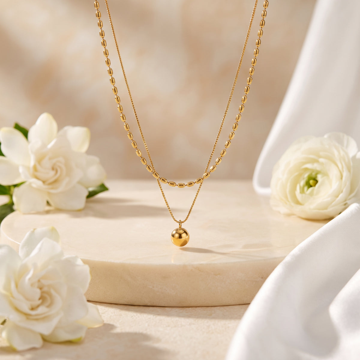 Gold-toned double-strand layered necklace suspended above a beige pedestal with white flowers.