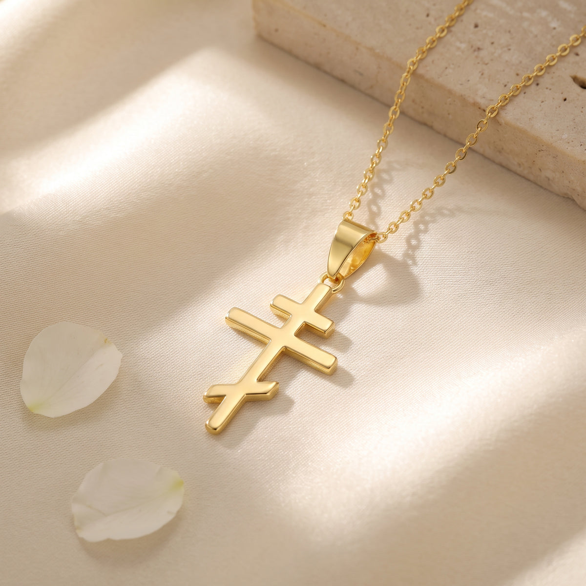 Gold-tone Eastern cross pendant necklace arranged on cream fabric beside white flower petals.