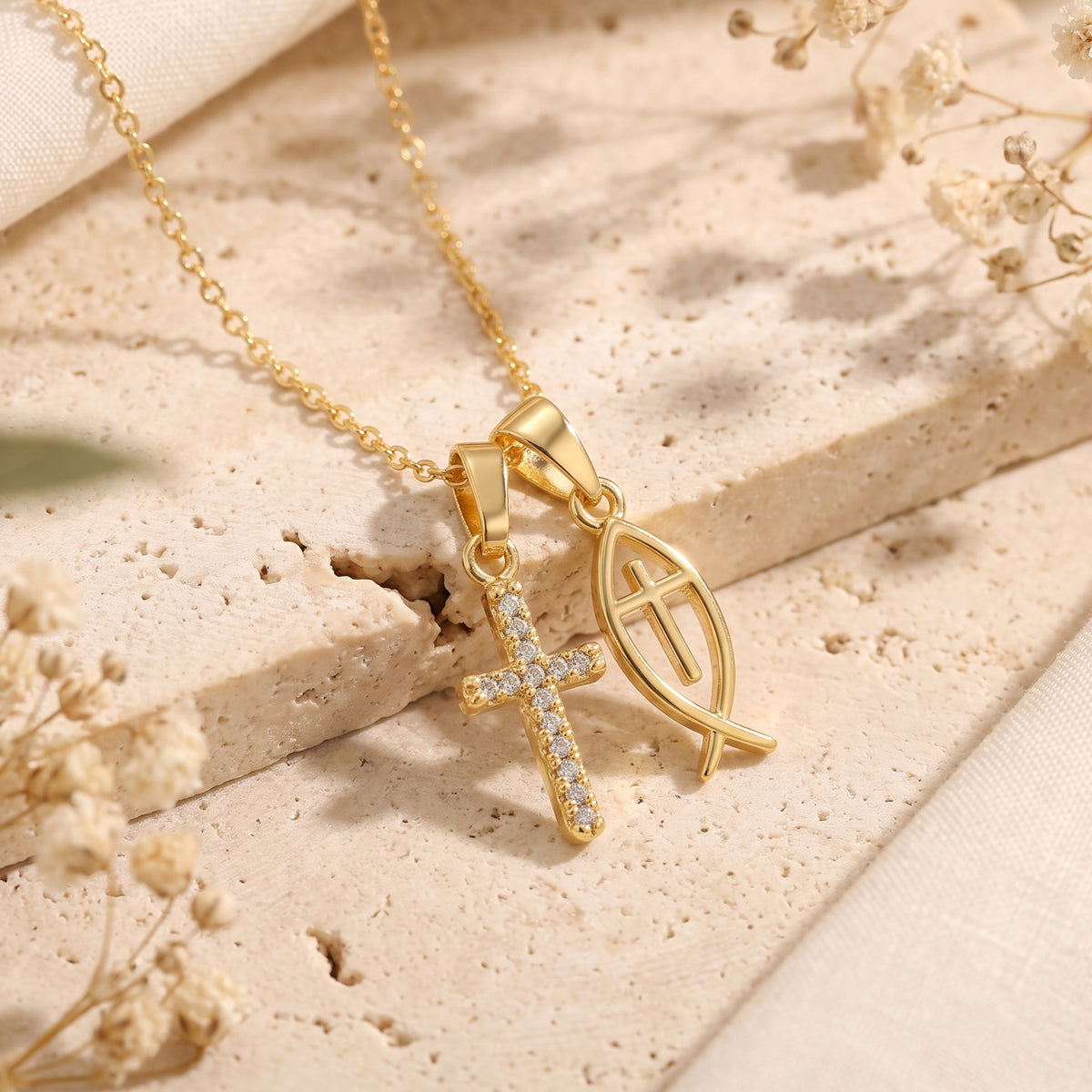 Close-up view of gold-tone necklace with cubic zirconia cross and Ichthys fish pendants on beige stone.