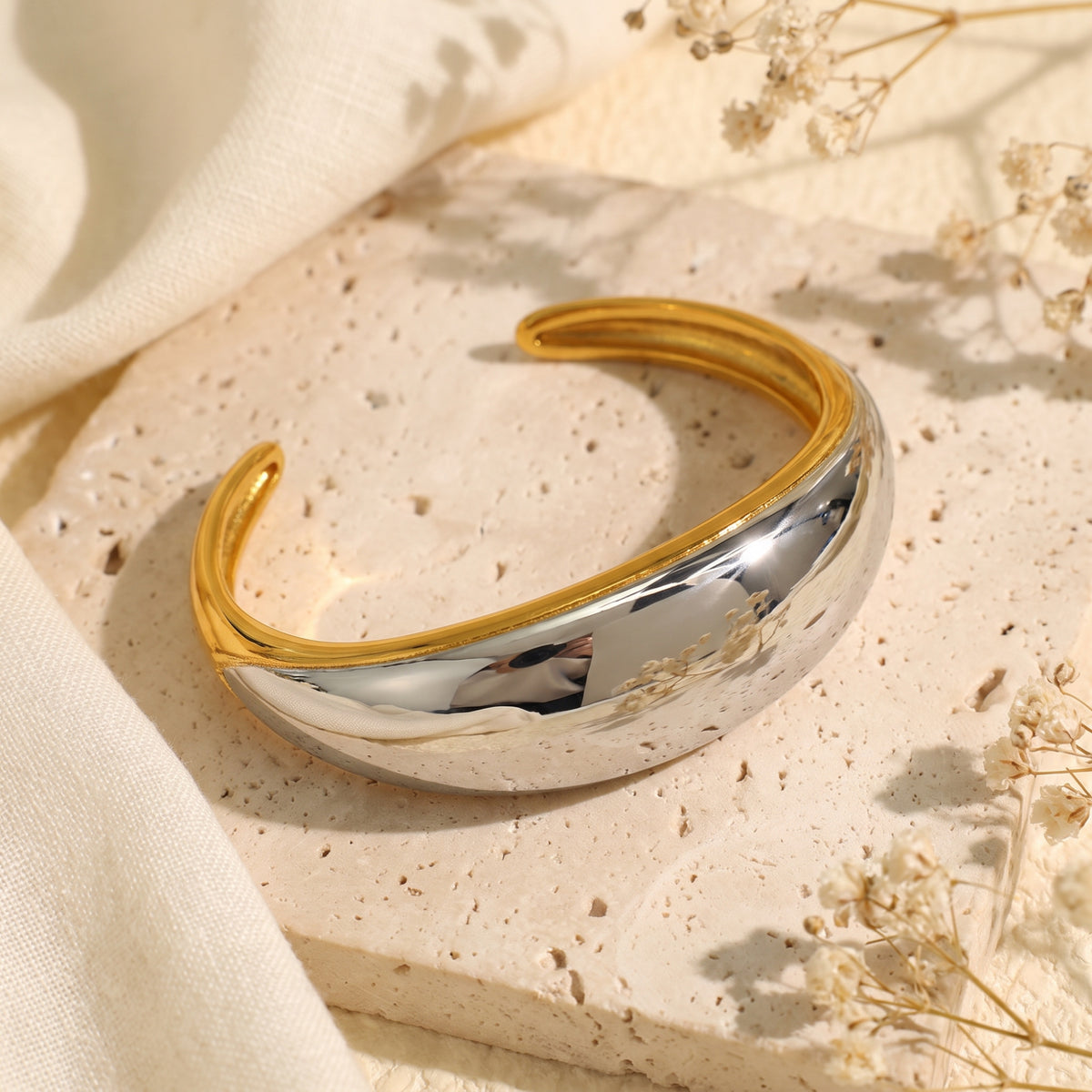 Dual-tone open cuff bracelet with a polished silver dome and gold-tone rim, resting on a beige stone slab.