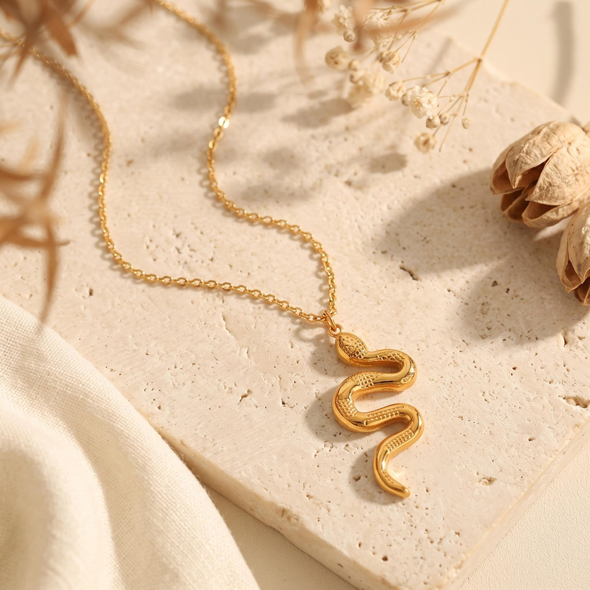 Gold-tone serpent pendant necklace resting on a beige stone surface beside dried botanical accents.