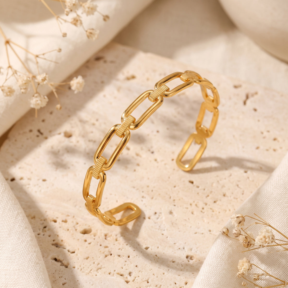 Gold-tone paperclip link open-cuff bracelet resting on a textured stone surface with cream fabric and dried flowers.