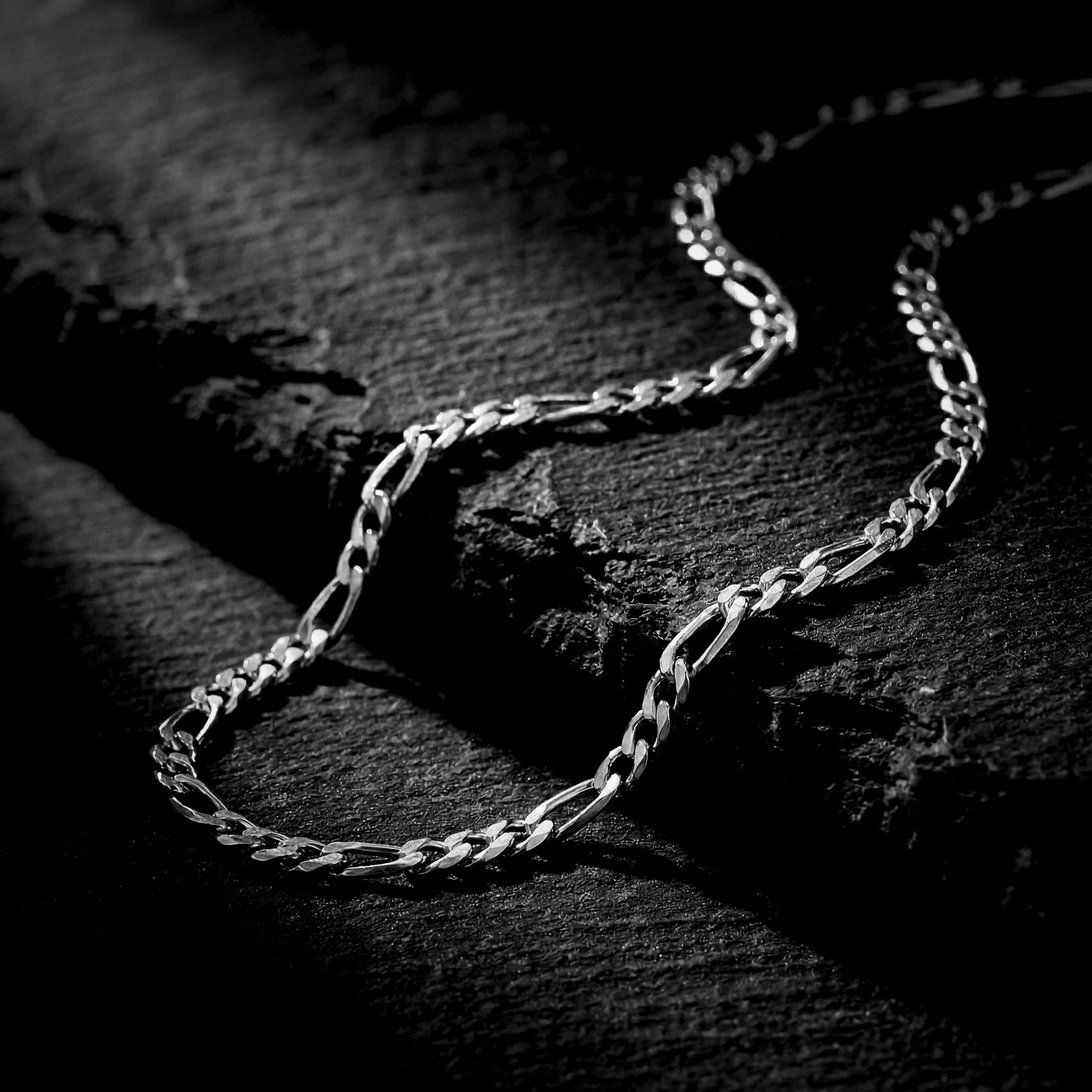 Close-up of a silver stainless steel 3mm Figaro chain on a dark stone background.