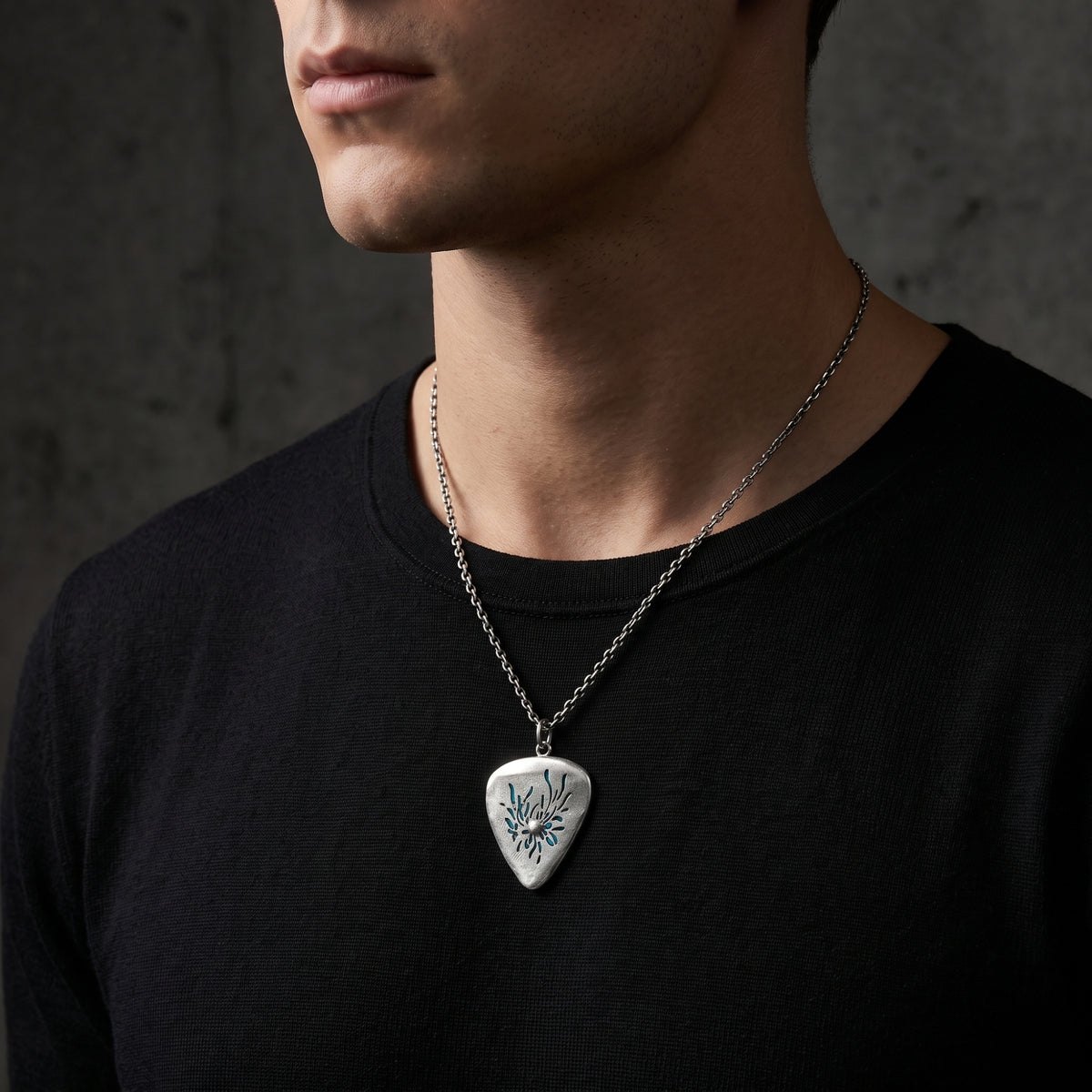 Man wearing the silver guitar-pick pendant necklace over a black crewneck shirt at mid-chest.