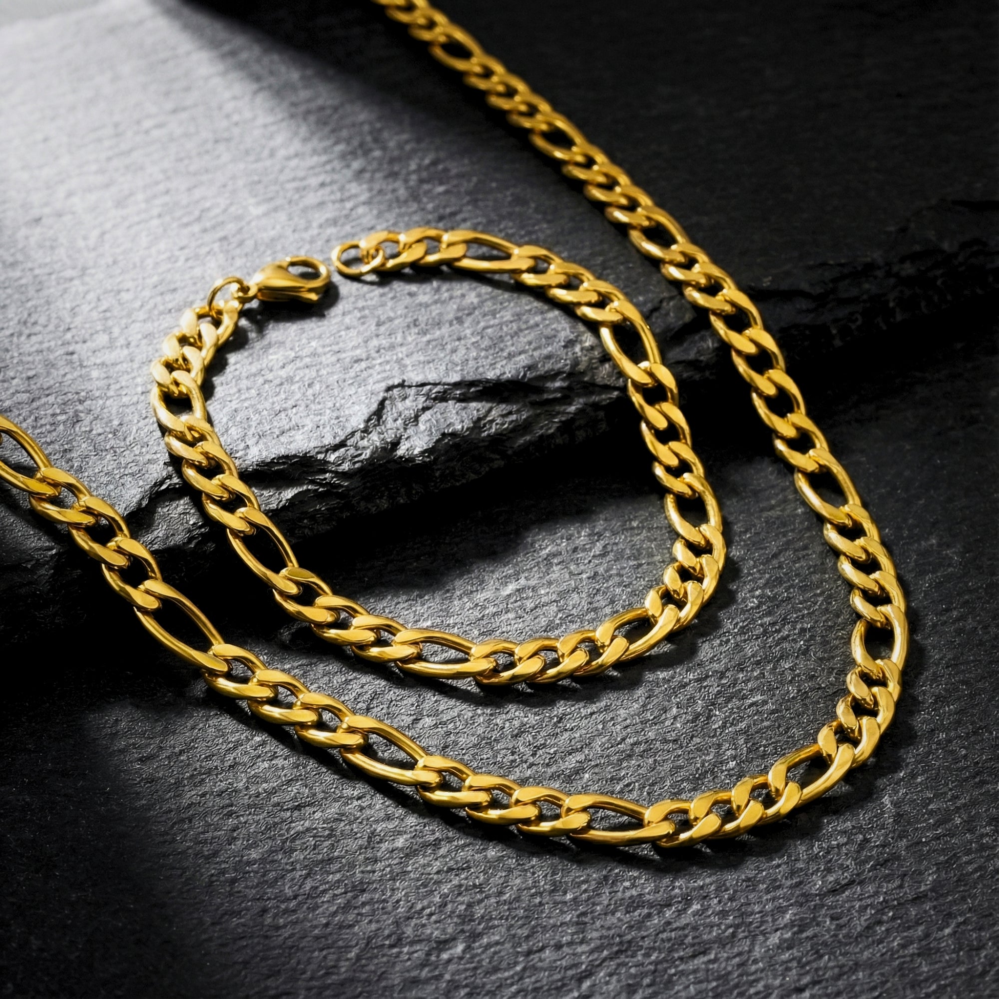 Close-up of a gold stainless steel Figaro chain and bracelet duo displayed on a textured black surface.