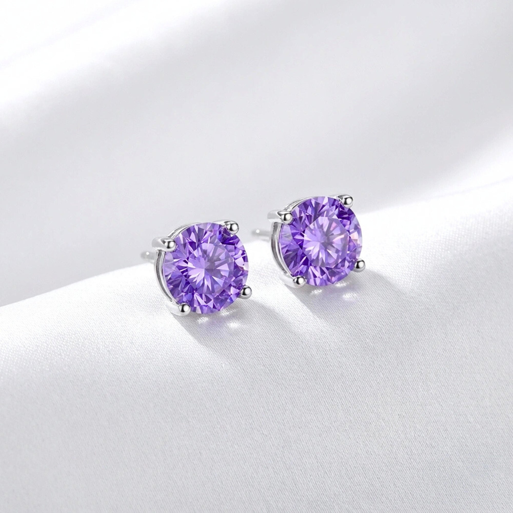 Side view of round cut simulated amethyst sterling silver stud earrings resting on white silk fabric.