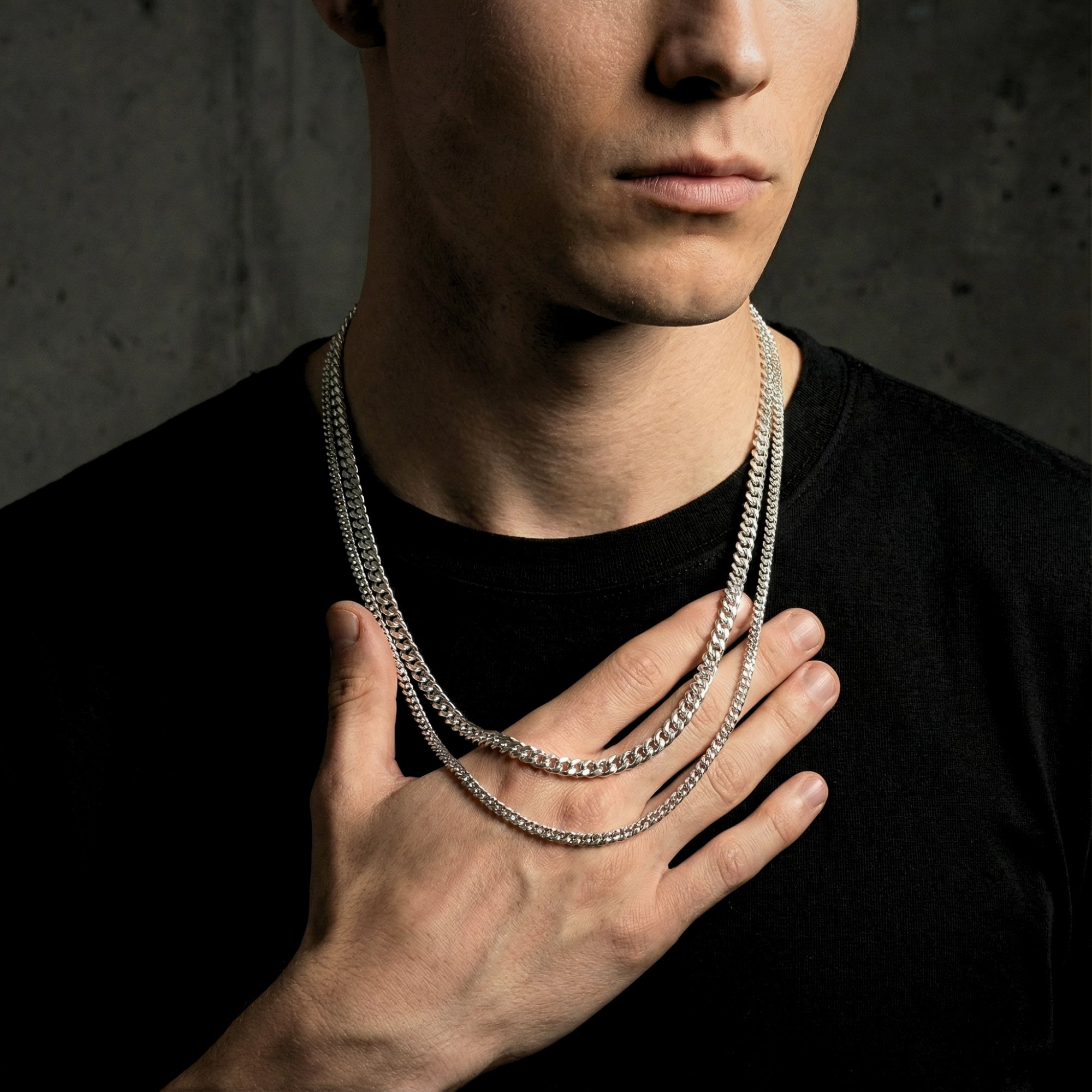 A man wearing the polished silver stainless steel Cuban Duo stacked chains, touching the necklaces with his hand.