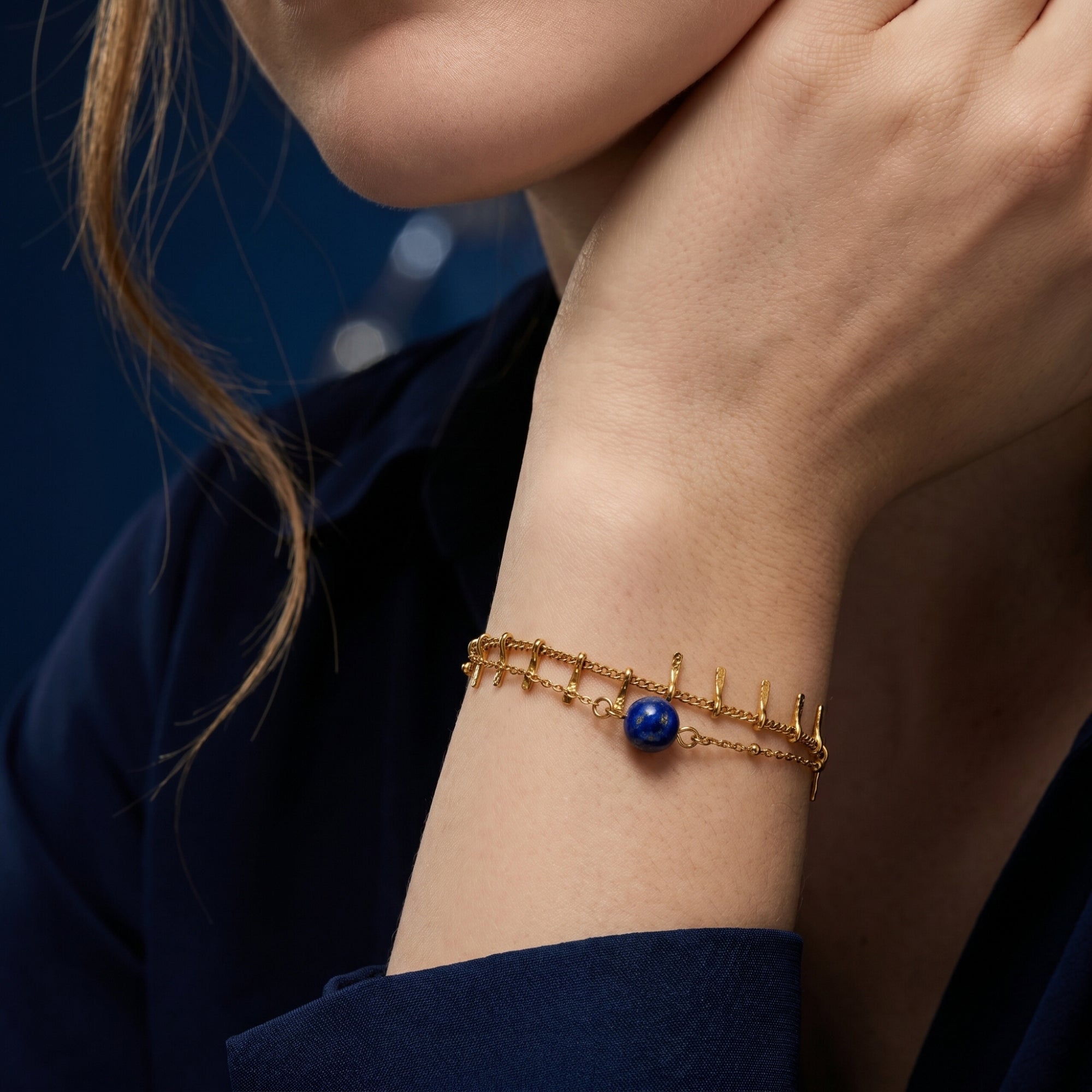 Close-up of a round blue lapis lazuli bead on a double-strand 18K gold plated chain bracelet on a wrist.